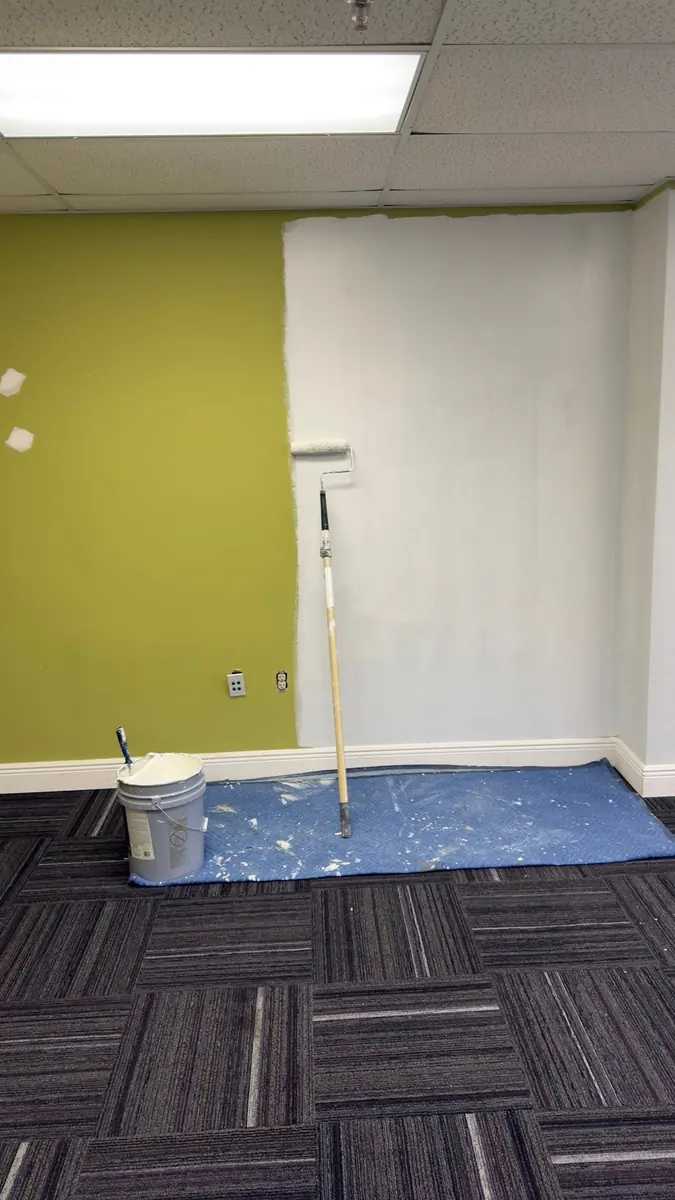 Office wall being repainted from green to white with a roller, with the carpet protected by a drop cloth