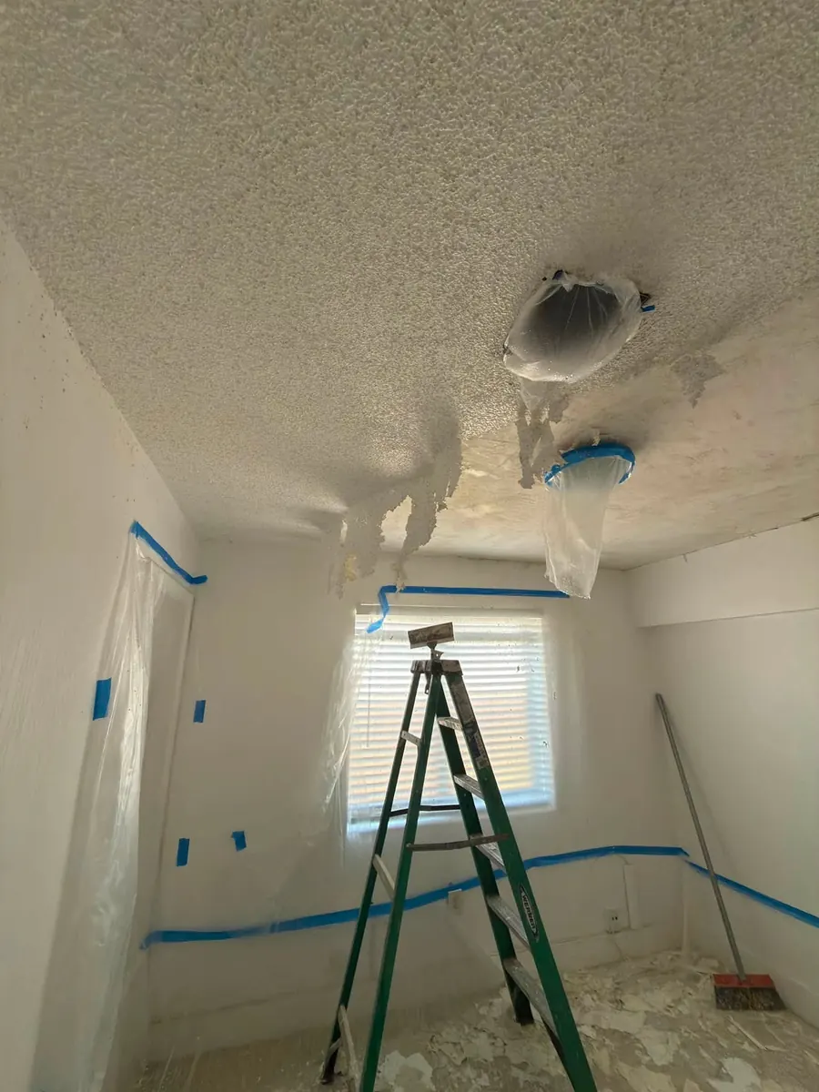 Popcorn ceiling texture being scraped off a bedroom ceiling, with light fixtures bagged and walls covered in plastic